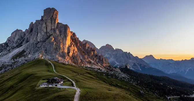 Evening light on the Ra Gusela rock wall above Passo Giau, with the pass hut and a gravel path on the green ridge below