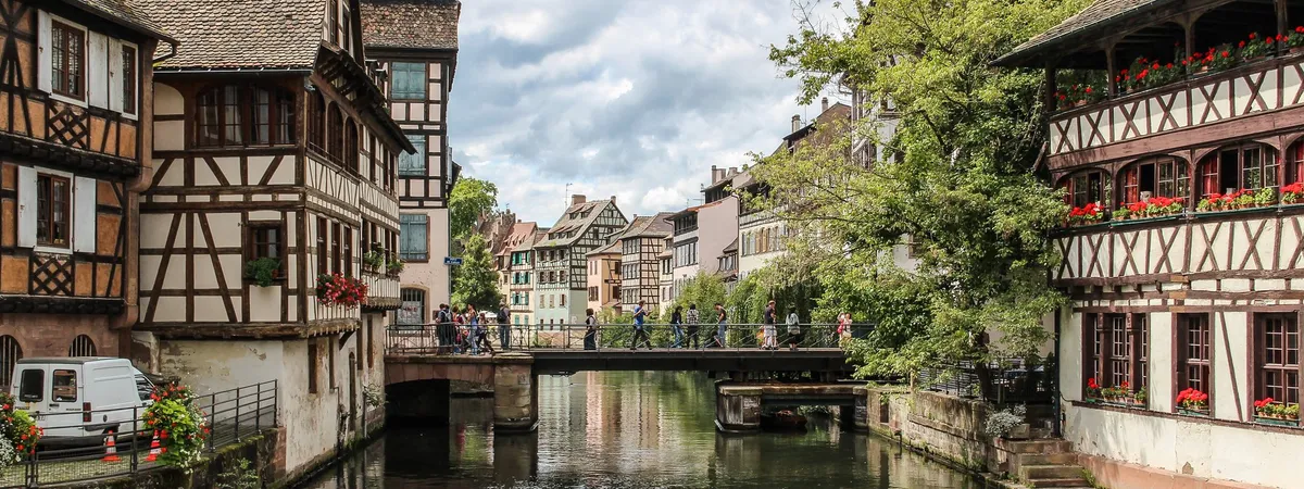 A canal in Strasbourg's Petite France quarter lined with half-timbered houses, a footbridge crossing the water, and geraniums on the balconies