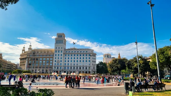 Multitudes y palomas llenando la Plaça de Catalunya, con el emblemático edificio del Banco Español de Crédito al fondo