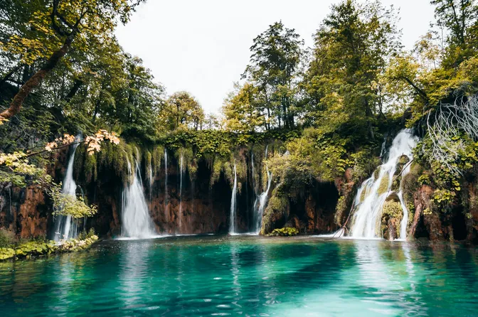 A row of waterfalls spilling over a mossy travertine ledge into a clear turquoise lake at Plitvice