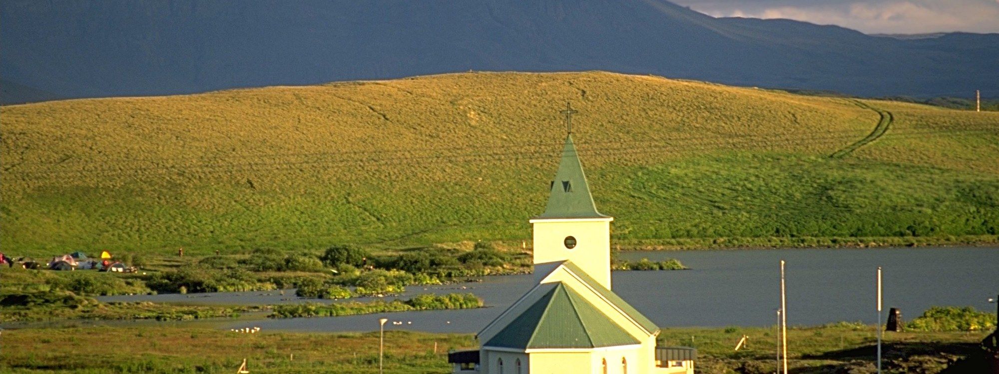 Reykjahlíð's white church with a green steeple, lit in golden evening light beside the shore of Lake Mývatn with mountains behind