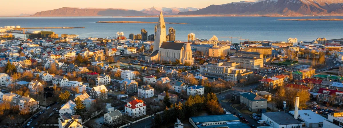 Aerial view of Reykjavík at sunset, with Hallgrímskirkja's tower rising above the city, Faxaflói bay, and snow-capped mountains beyond