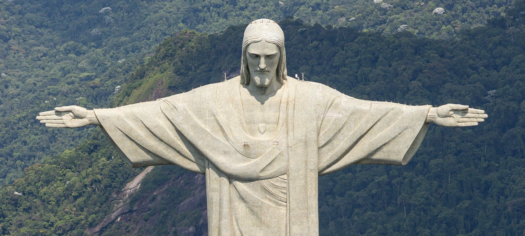 The outstretched arms of the Christ the Redeemer statue atop Corcovado mountain, seen close up against the forested hillside