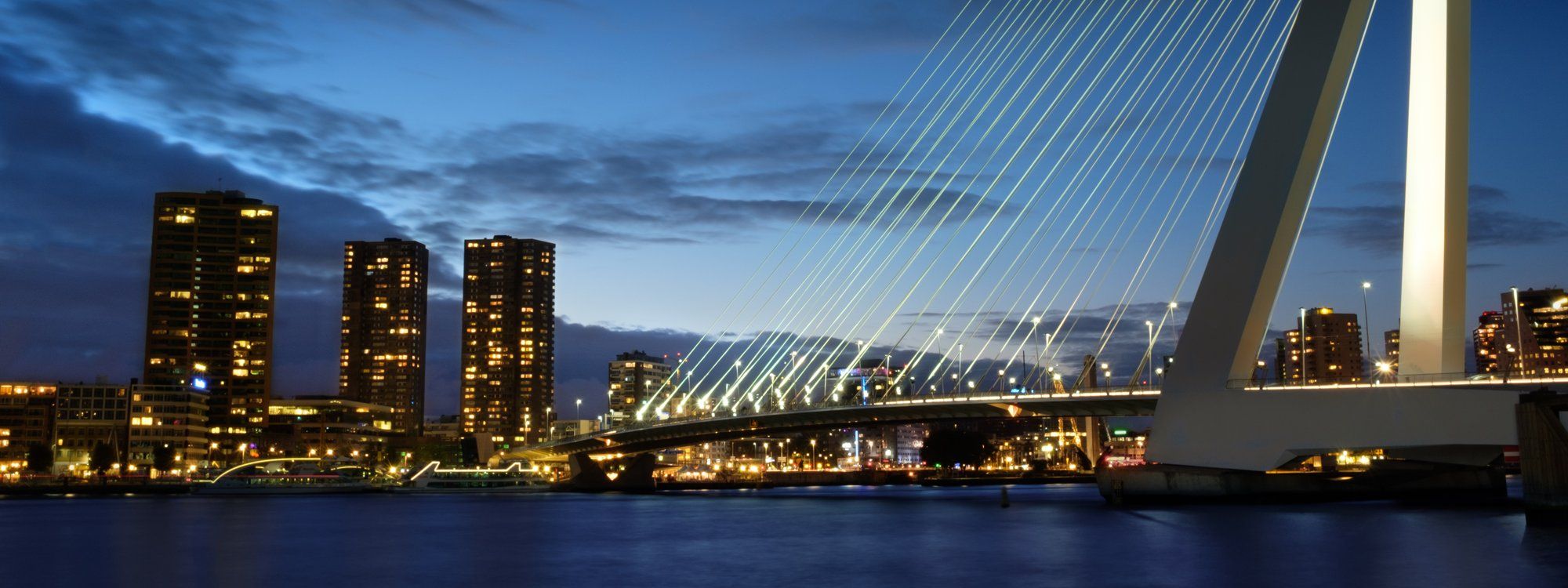 The Erasmus Bridge lit up at night alongside the Rotterdam skyline, reflected in the Nieuwe Maas river