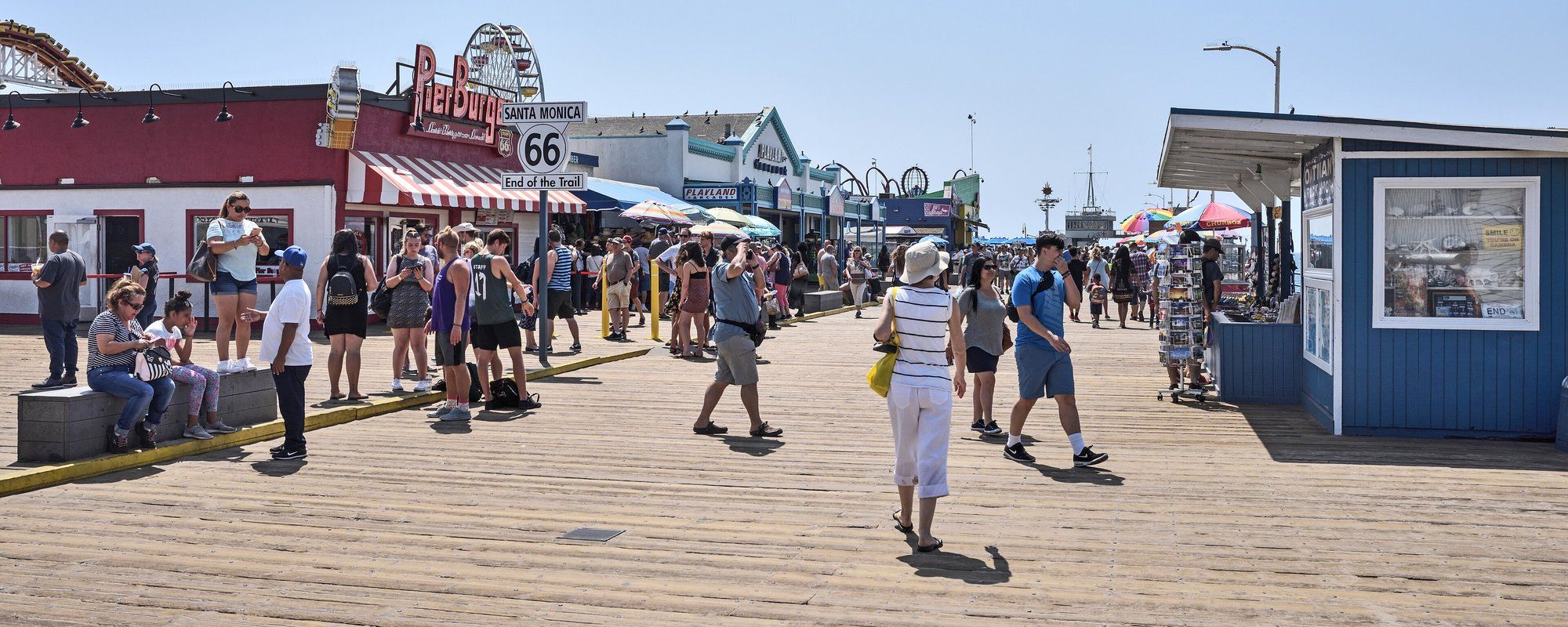 Crowds walking the wooden deck of Santa Monica Pier past the Route 66 "End of the Trail" sign, with the pier's carnival rides behind them