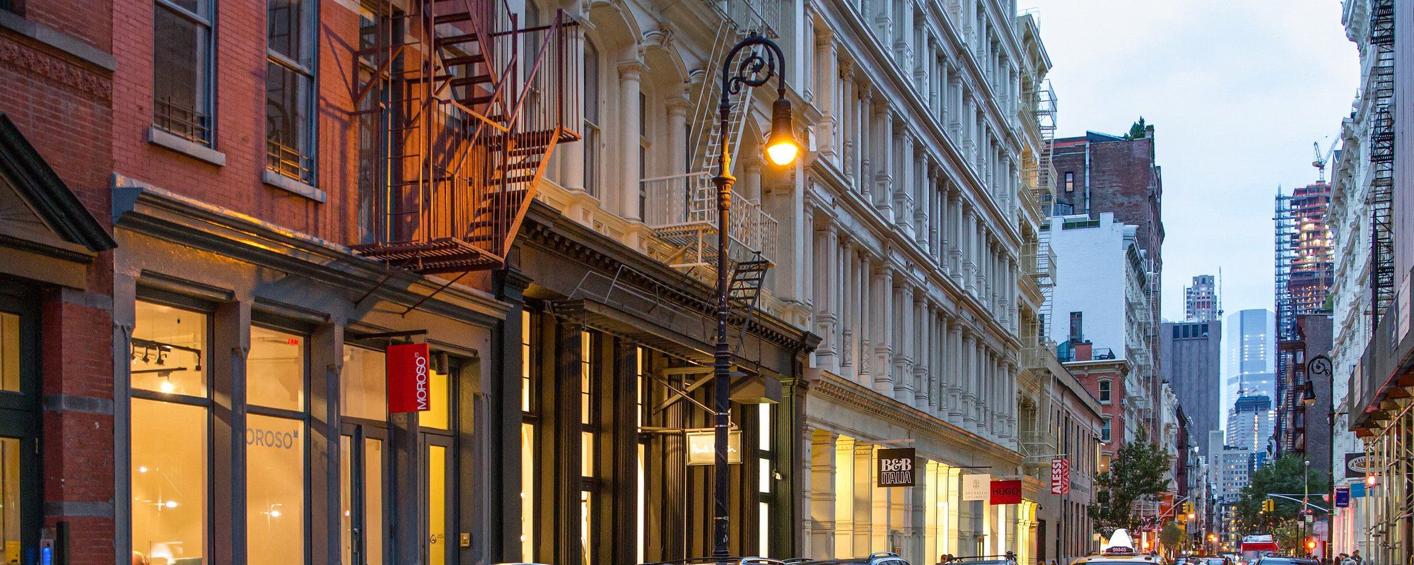 Greene Street in SoHo at dusk, lined with 19th-century cast-iron building facades, fire escapes, and lit storefronts