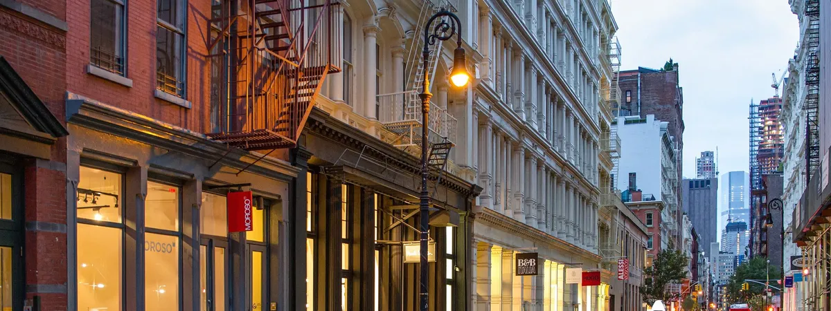 Greene Street in SoHo at dusk, lined with 19th-century cast-iron building facades, fire escapes, and lit storefronts