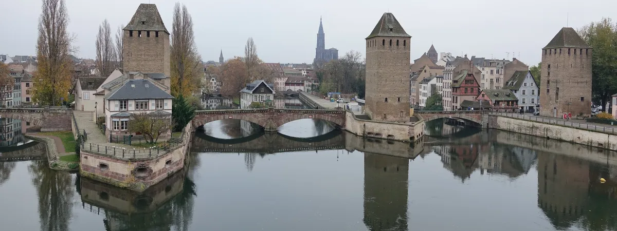 The medieval Ponts Couverts towers and Petite France's half-timbered houses reflected in the Ill river, with Strasbourg Cathedral's spire visible on the skyline behind