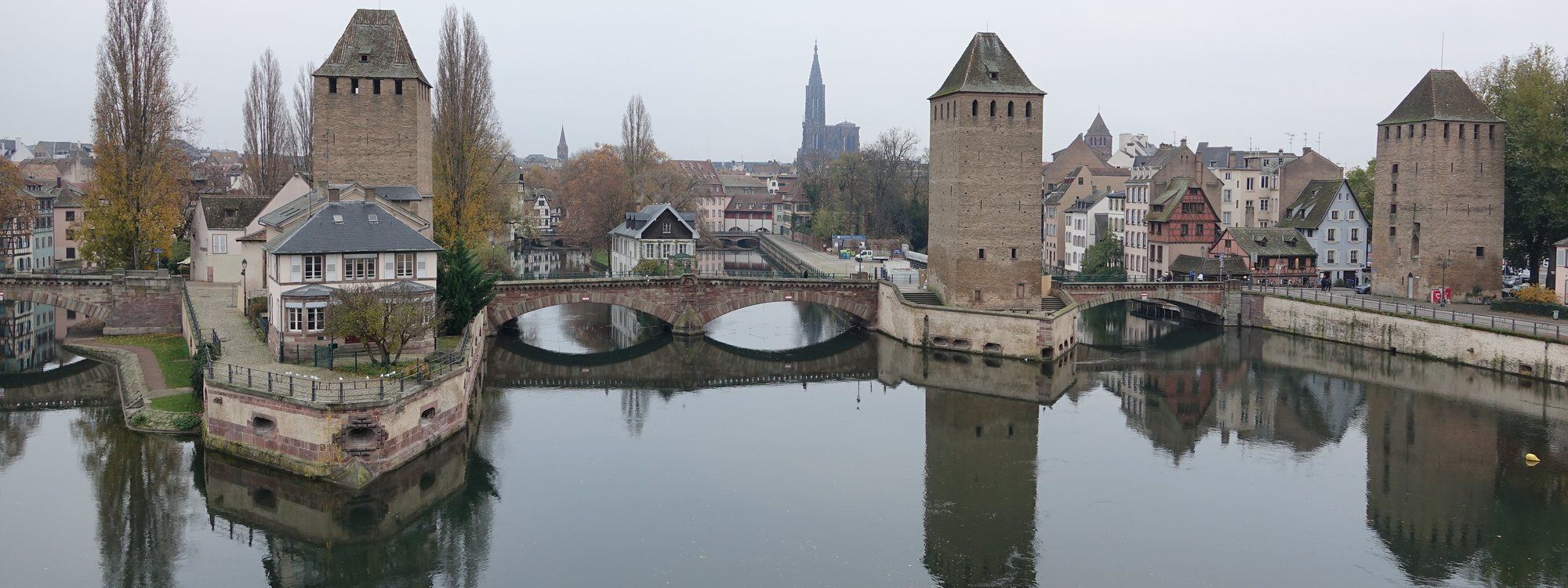 The medieval Ponts Couverts towers and Petite France's half-timbered houses reflected in the Ill river, with Strasbourg Cathedral's spire visible on the skyline behind