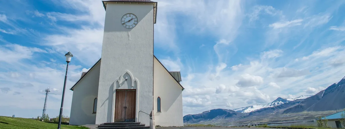 Borgarneskirkja, the white church in Borgarnes, standing against a blue sky with snow-capped mountains across the fjord behind it