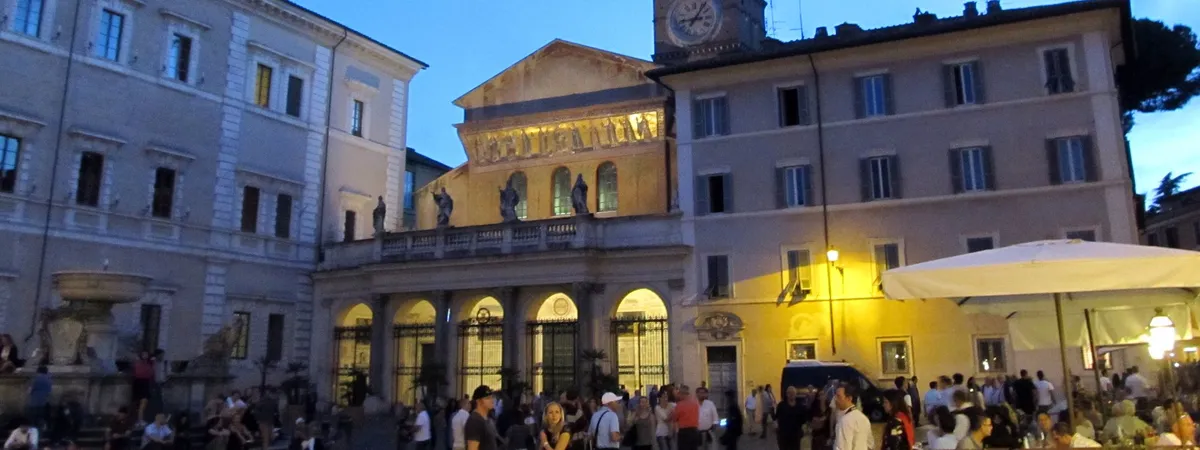 The gold mosaic facade of the Basilica of Santa Maria in Trastevere lit up at dusk, with the piazza in front of it full of people