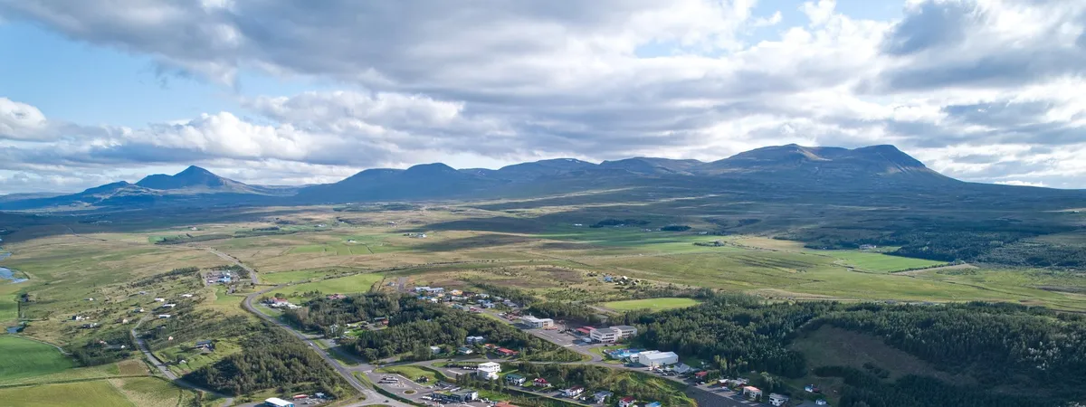 Aerial view of Varmahlíð village on the floor of the Skagafjörður valley, with a forested hill and farmland around it and mountains behind