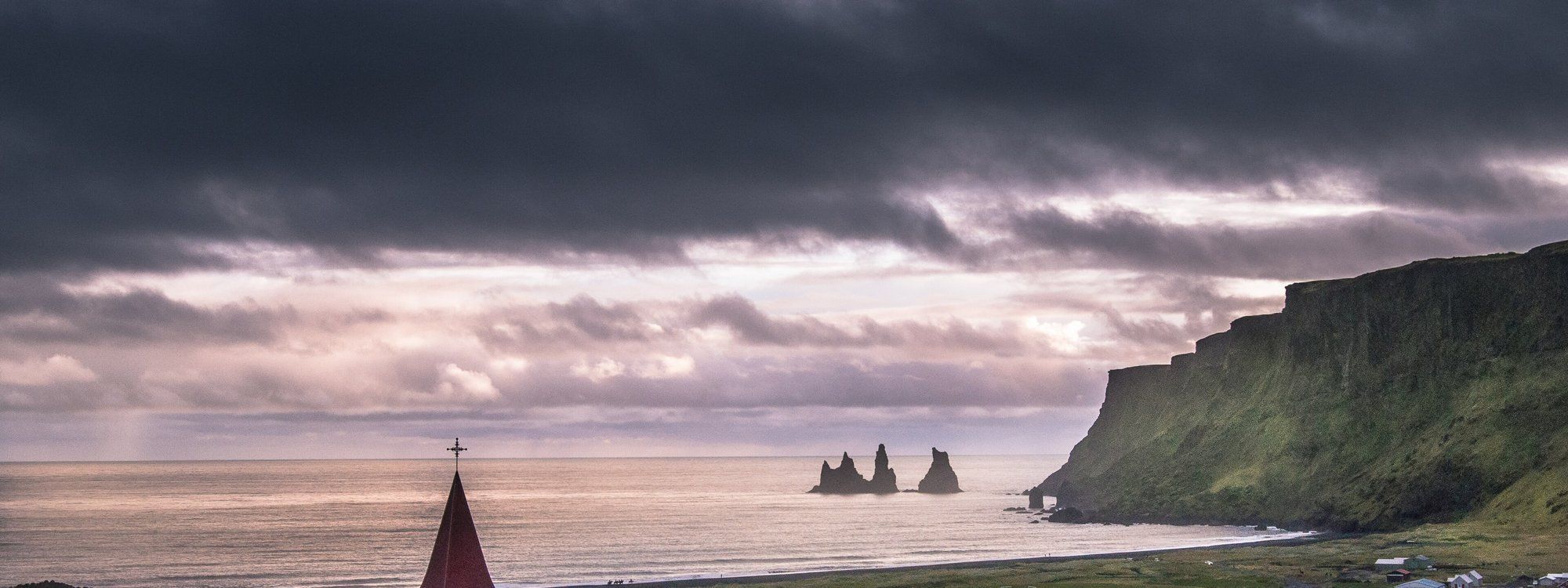 Víkurkirkja's red spire overlooking Vík's black-sand coastline and the Reynisdrangar sea stacks under a dramatic sky