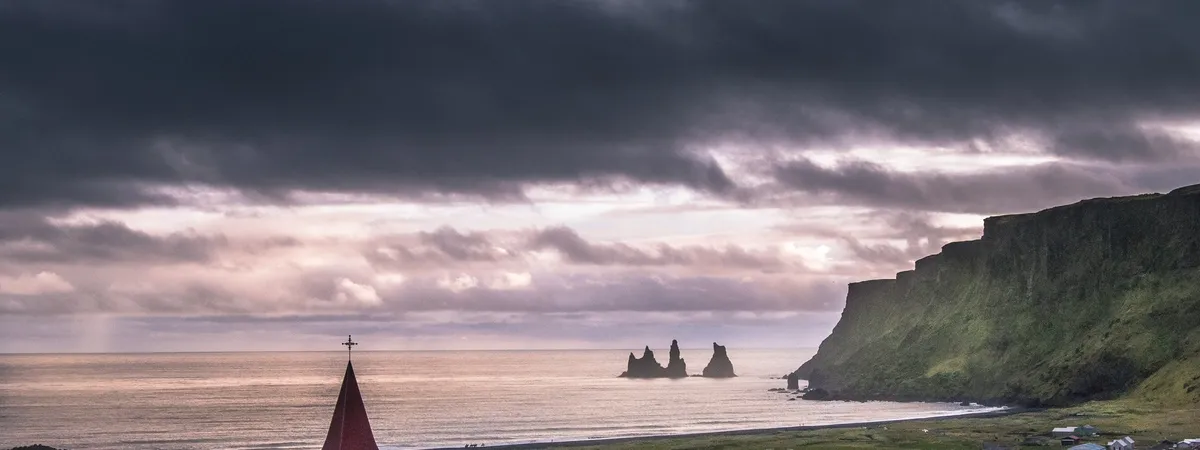 Víkurkirkja's red spire overlooking Vík's black-sand coastline and the Reynisdrangar sea stacks under a dramatic sky