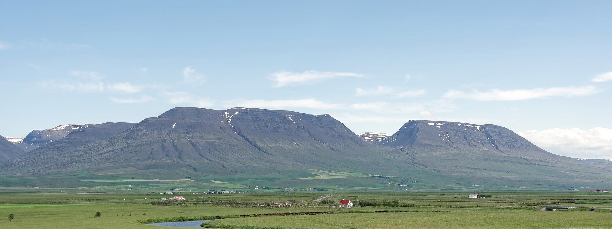 The Svartá river winding through green farmland beside Varmahlíð, with a red-roofed farmhouse and layered mountains behind