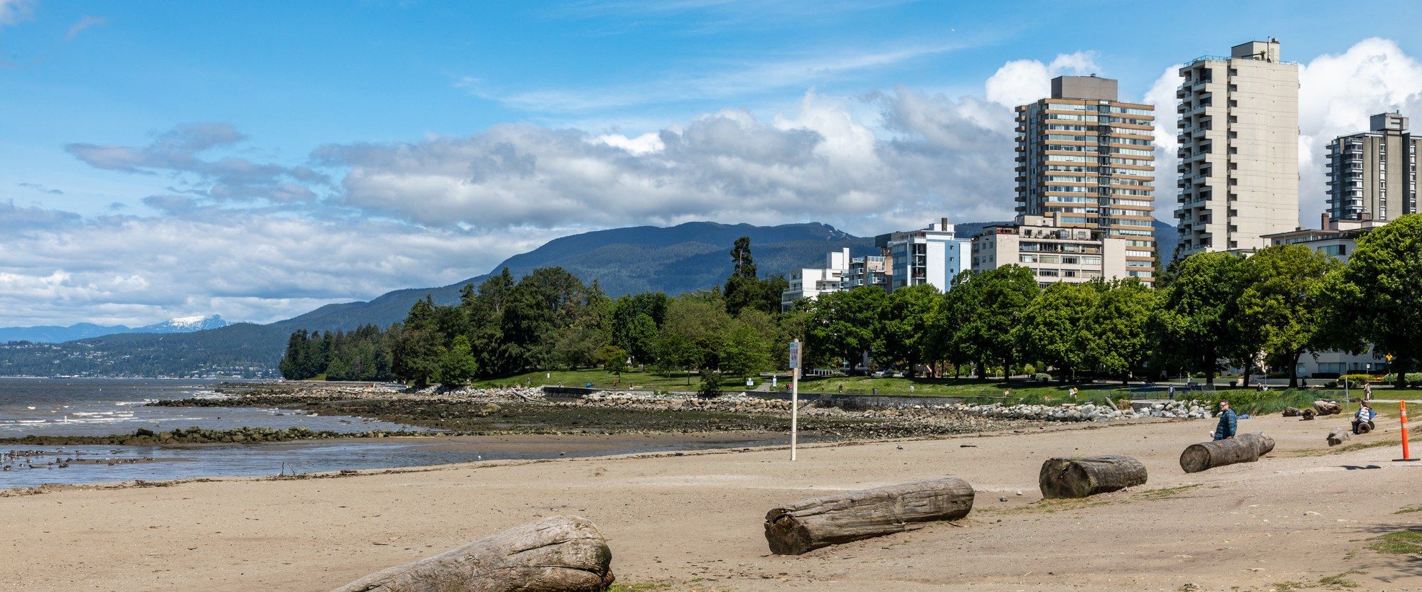 English Bay Beach in Vancouver's West End, with driftwood logs on the sand, apartment towers, and the North Shore mountains behind