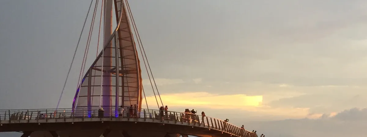The sail-shaped pavilion of Los Muertos Pier at sunset, with people gathered along the railing and small boats anchored in the water below